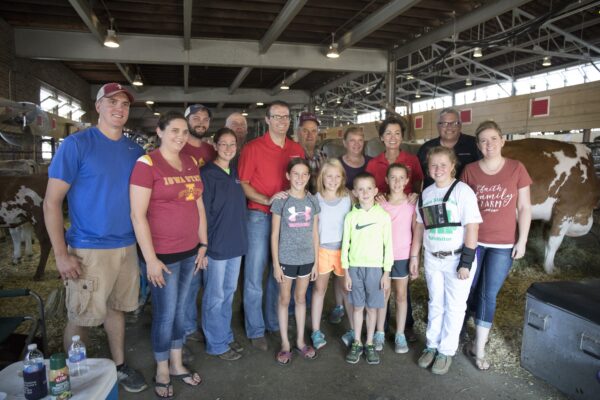 Group of children and adults stand in a barn with cows.