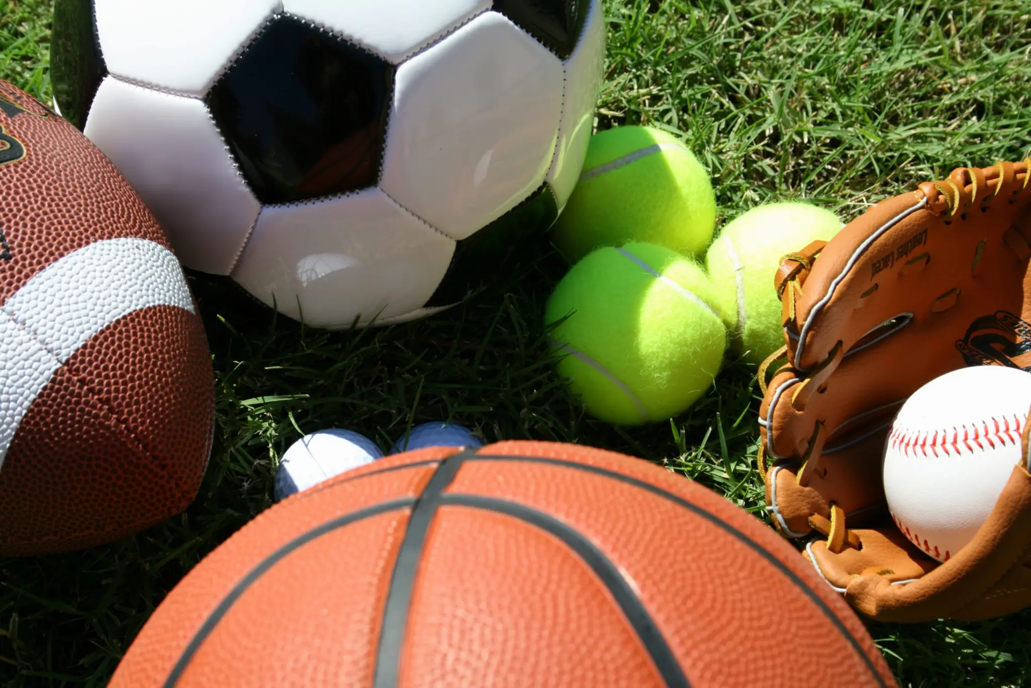 A football, soccer ball, basketball, three tennis balls, two golf balls, and a baseball in a leather baseball glove on grass.