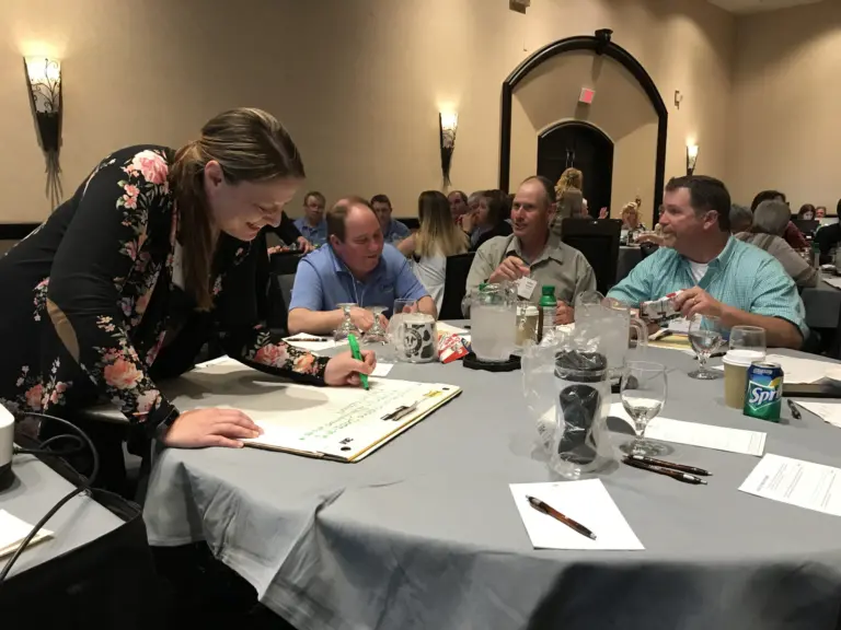 A woman writes on a large clipboard while three men sit at a table.