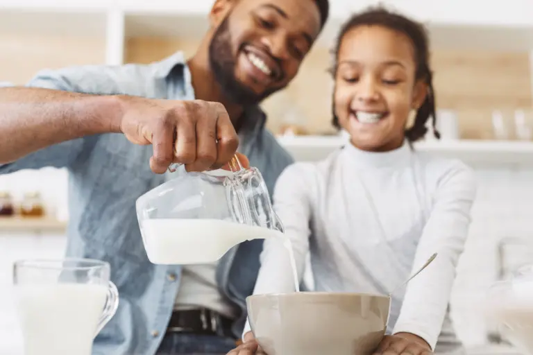 Happy man pouring milk into daughter's cereals, having breakfast together