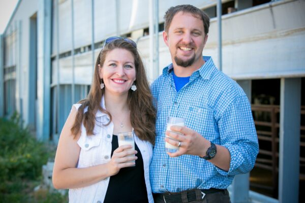 A couple poses in front of a barn, holding glasses of milk.