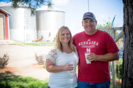 Man and woman standing on a farm holding glasses of milk with steel grain bins in the background.