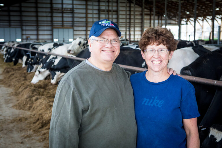 Middle-aged couple stands in a barn, with cows eating in the background.