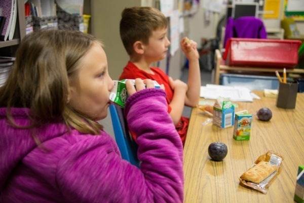 A boy and a girl eating a snack and drinking milk at a table in a classroom.