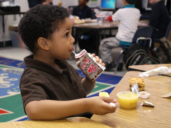 A little boy seated at a table in the school cafeteria, drinking a carton of chocolate milk.
